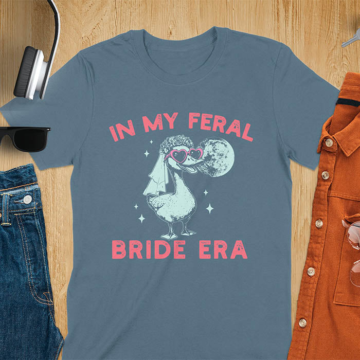 A goose standing, wearing heart-shaped sunglasses and a bride's veil. Moon in the upper right background. Retro-style text reads In My Feral Bride Era and Bride Era. Whimsical image.
