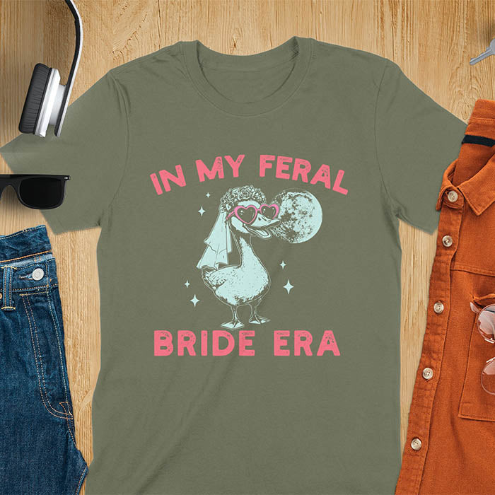 A goose standing, wearing heart-shaped sunglasses and a bride's veil. Moon in the upper right background. Retro-style text reads In My Feral Bride Era and Bride Era. Whimsical image.
