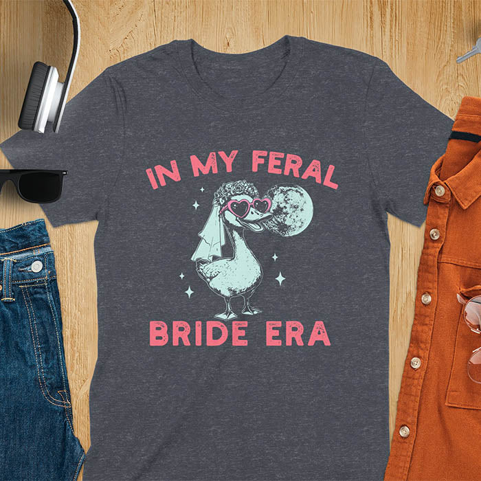 A goose standing, wearing heart-shaped sunglasses and a bride's veil. Moon in the upper right background. Retro-style text reads In My Feral Bride Era and Bride Era. Whimsical image.