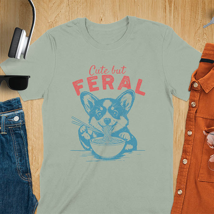 A t-shirt with a corgi eating noodles with chopsticks. Text: Cute but FERAL above. This funny dog shirt features kawaiiness cuteness, a meme graphic tee. A retro dog shirt and vintage dog tee, an animal person gift or gift mom hers. A funny unhinge shirt or weird funny shirt for a dog dad shirt. A comfortable t-shirts option with a trendy tshirt design, a funny animal meme.