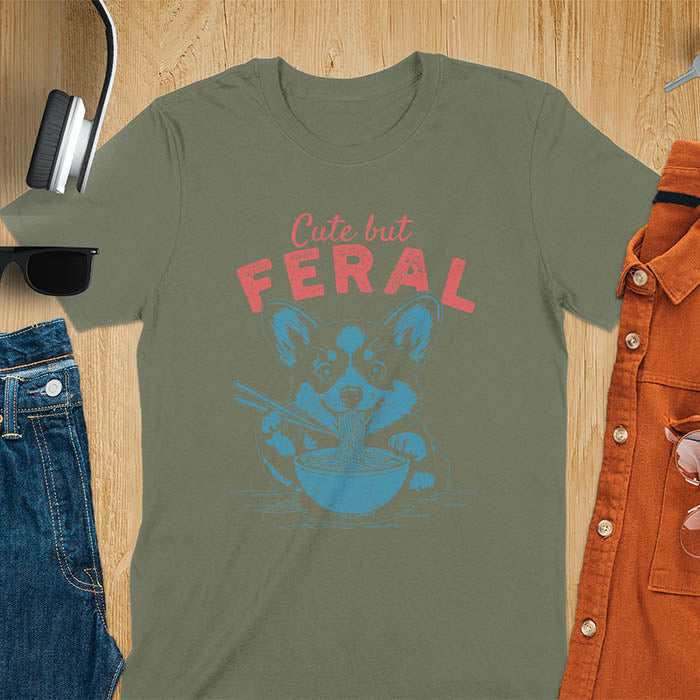 A t-shirt with a corgi eating noodles with chopsticks. Text: Cute but FERAL above. This funny dog shirt features kawaiiness cuteness, a meme graphic tee. A retro dog shirt and vintage dog tee, an animal person gift or gift mom hers. A funny unhinge shirt or weird funny shirt for a dog dad shirt. A comfortable t-shirts option with a trendy tshirt design, a funny animal meme.