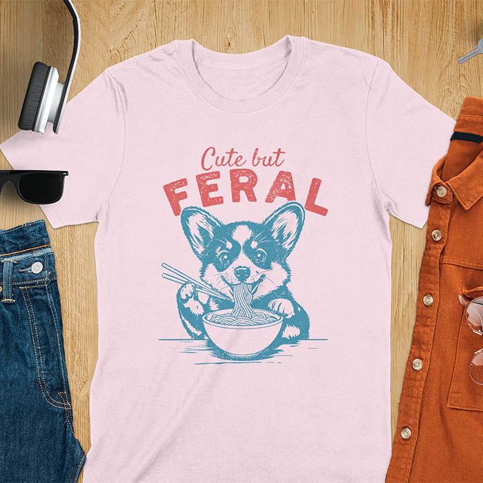 A t-shirt with a corgi eating noodles with chopsticks. Text: Cute but FERAL above. This funny dog shirt features kawaiiness cuteness, a meme graphic tee. A retro dog shirt and vintage dog tee, an animal person gift or gift mom hers. A funny unhinge shirt or weird funny shirt for a dog dad shirt. A comfortable t-shirts option with a trendy tshirt design, a funny animal meme.