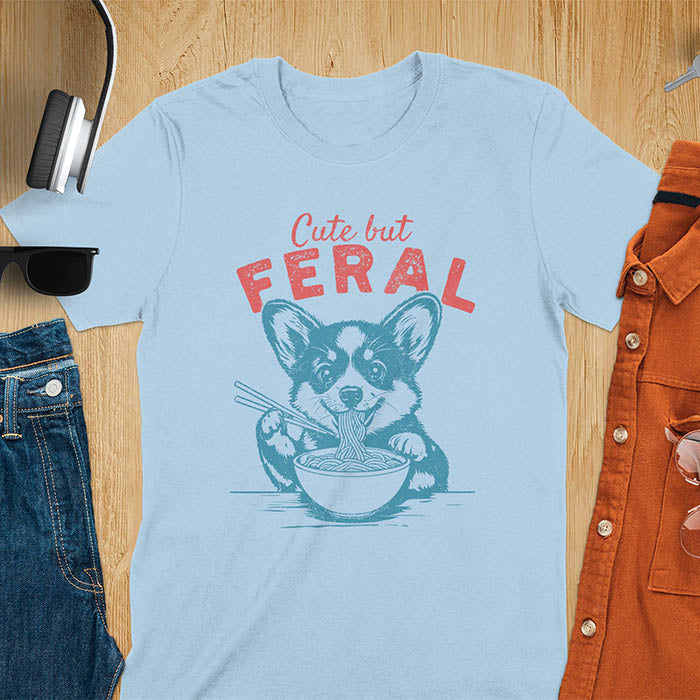 A t-shirt with a corgi eating noodles with chopsticks. Text: Cute but FERAL above. This funny dog shirt features kawaiiness cuteness, a meme graphic tee. A retro dog shirt and vintage dog tee, an animal person gift or gift mom hers. A funny unhinge shirt or weird funny shirt for a dog dad shirt. A comfortable t-shirts option with a trendy tshirt design, a funny animal meme.
