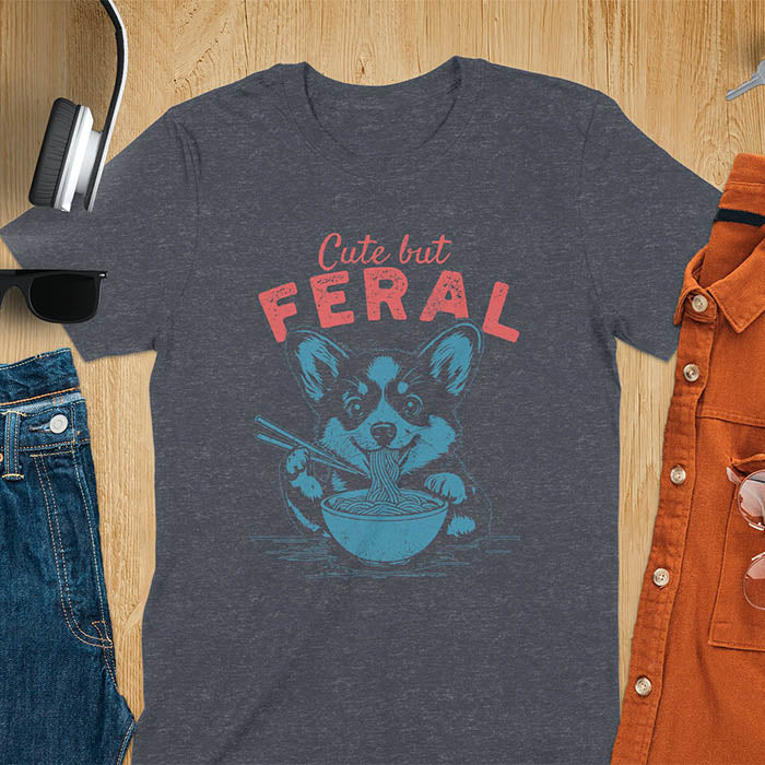 A t-shirt with a corgi eating noodles with chopsticks. Text: Cute but FERAL above. This funny dog shirt features kawaiiness cuteness, a meme graphic tee. A retro dog shirt and vintage dog tee, an animal person gift or gift mom hers. A funny unhinge shirt or weird funny shirt for a dog dad shirt. A comfortable t-shirts option with a trendy tshirt design, a funny animal meme.