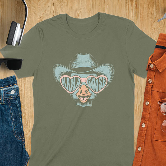 A t-shirt featuring a goose wearing a cowboy hat and oversized pink heart-shaped sunglasses. The pink lenses of the sunglasses display the words Mama Goose in a stylized font. The goose graphic is centered on the shirt and has a vintage distressed texture.