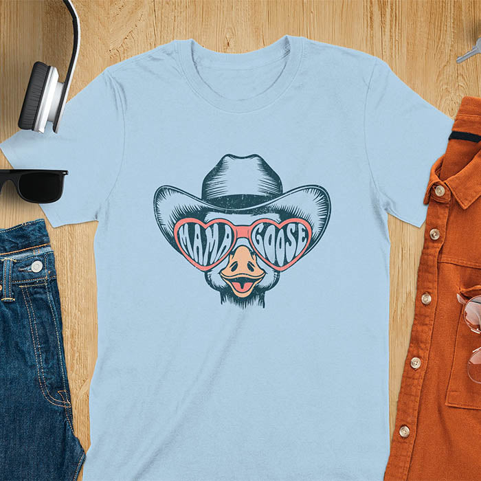 A t-shirt featuring a goose wearing a cowboy hat and oversized pink heart-shaped sunglasses. The pink lenses of the sunglasses display the words Mama Goose in a stylized font. The goose graphic is centered on the shirt and has a vintage distressed texture.