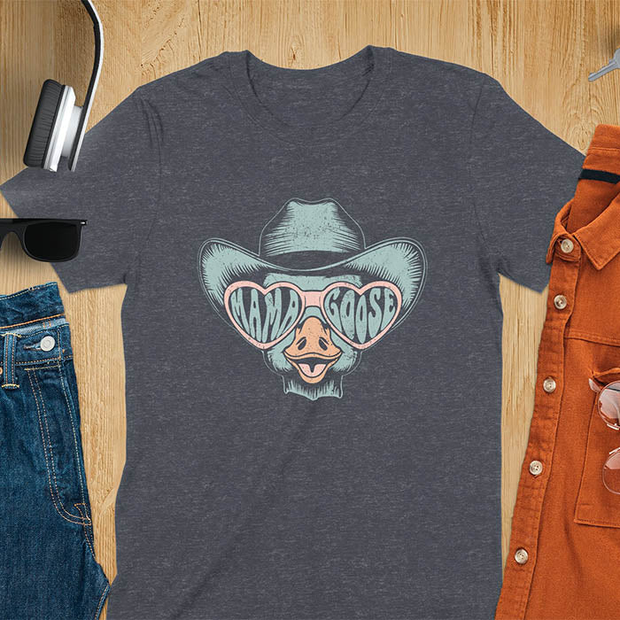 A t-shirt featuring a goose wearing a cowboy hat and oversized pink heart-shaped sunglasses. The pink lenses of the sunglasses display the words Mama Goose in a stylized font. The goose graphic is centered on the shirt and has a vintage distressed texture.