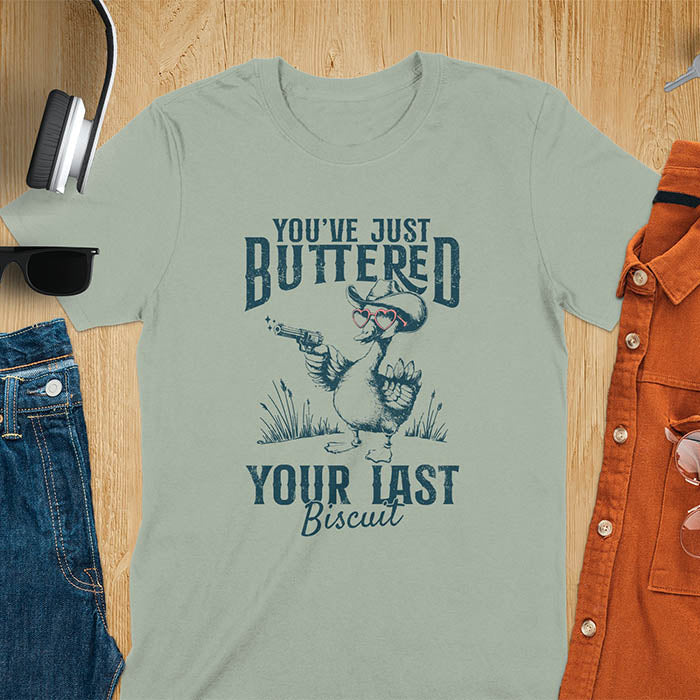 A t-shirt with a goose wearing a cowboy hat and sunglasses, holding a pistol. Text: "YOU'VE JUST BUTTERED" above and "YOUR LAST Biscuit" below. This funny cowboy shirt features a silly goose shirt or goose lovers gift. A cowgirl t-shirt and country girl shirt, a western girl shirt. A retro graphic tee and weirdcore tee-shirt, an animal person gift or animal lover gag. A mom gift alternative, a last biscuit shirt with a sarcastic quote.