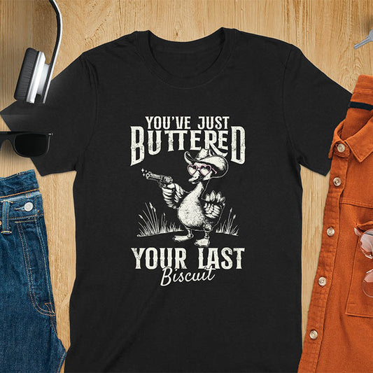 A t-shirt with a goose wearing a cowboy hat and sunglasses, holding a pistol. Text: "YOU'VE JUST BUTTERED" above and "YOUR LAST Biscuit" below. This funny cowboy shirt features a silly goose shirt or goose lovers gift. A cowgirl t-shirt and country girl shirt, a western girl shirt. A retro graphic tee and weirdcore tee-shirt, an animal person gift or animal lover gag. A mom gift alternative, a last biscuit shirt with a sarcastic quote.