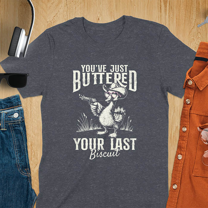A t-shirt with a goose wearing a cowboy hat and sunglasses, holding a pistol. Text: "YOU'VE JUST BUTTERED" above and "YOUR LAST Biscuit" below. This funny cowboy shirt features a silly goose shirt or goose lovers gift. A cowgirl t-shirt and country girl shirt, a western girl shirt. A retro graphic tee and weirdcore tee-shirt, an animal person gift or animal lover gag. A mom gift alternative, a last biscuit shirt with a sarcastic quote.