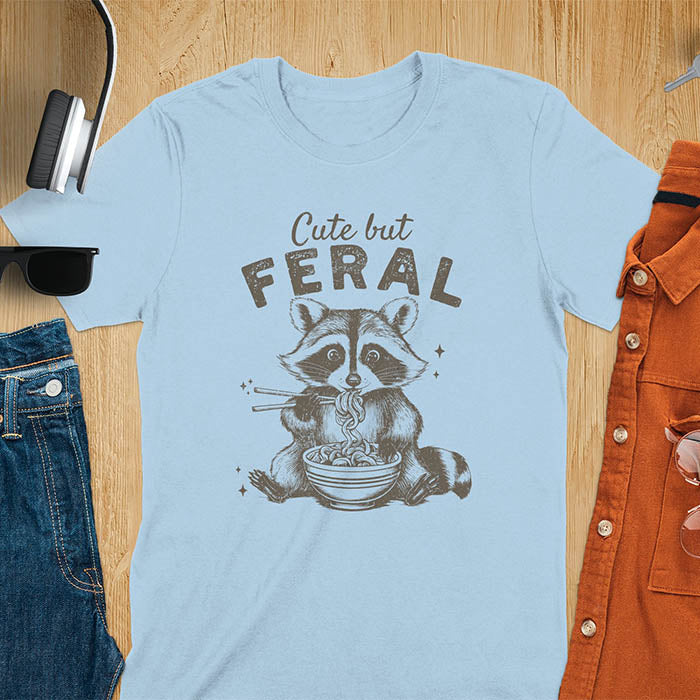 A t-shirt with a raccoon eating ramen with chopsticks. Text above: "Cute but". Text below: "FERAL". This Cute But Feral Shirt is a Funny Raccoon T-shirt, Vintage Retro Graphic Ramen Tee-shirt. A Silly Meme Tee, Hilarious Gag Gift, funny unhinge shirt, sarcastic raccoon, trendy tshirt design. Features kawaiiness cuteness. Ideal for animal person gift, raccoon lover shirt, gift mom hers, feral mom t shirt. A vintage inspired Animal Lover Tshirt, almost feral shirt.