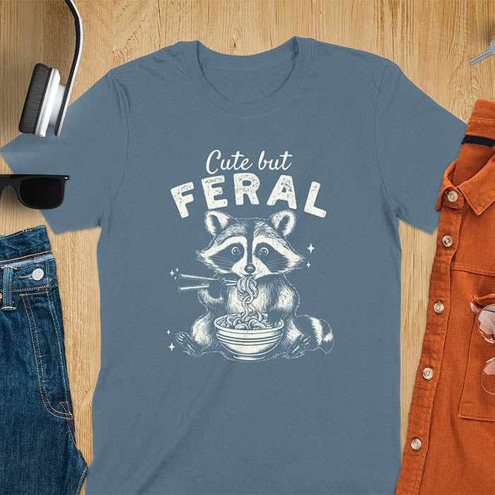 A t-shirt with a raccoon eating ramen with chopsticks. Text above: "Cute but". Text below: "FERAL". This Cute But Feral Shirt is a Funny Raccoon T-shirt, Vintage Retro Graphic Ramen Tee-shirt. A Silly Meme Tee, Hilarious Gag Gift, funny unhinge shirt, sarcastic raccoon, trendy tshirt design. Features kawaiiness cuteness. Ideal for animal person gift, raccoon lover shirt, gift mom hers, feral mom t shirt. A vintage inspired Animal Lover Tshirt, almost feral shirt.