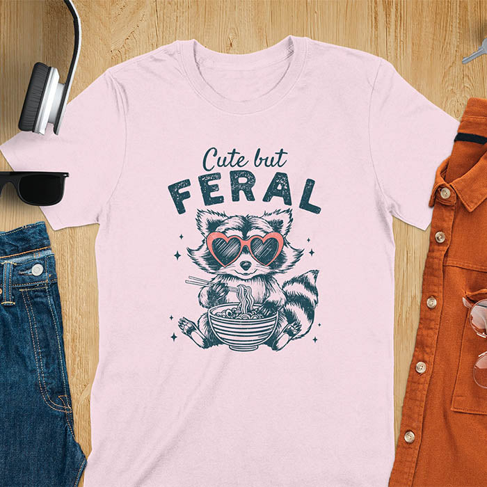 A t-shirt with a raccoon in heart sunglasses eating ramen. Text: "Cute but FERAL". A Funny Cute But Feral Raccoon Shirt, Vintage Retro Graphic Ramen Tee, Silly Sarcastic Tshirt, Hilarious Gag Gift. Unhinged, trash panda, trendy design. Features kawaiiness cuteness. Great gift for animal person, raccoon lover, mom, feral mom. Vintage inspired Animal Lover T-shirt, almost feral shirt.