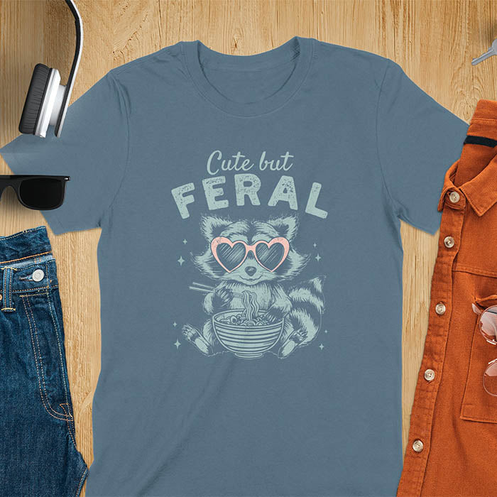 A t-shirt with a raccoon in heart sunglasses eating ramen. Text: "Cute but FERAL". A Funny Cute But Feral Raccoon Shirt, Vintage Retro Graphic Ramen Tee, Silly Sarcastic Tshirt, Hilarious Gag Gift. Unhinged, trash panda, trendy design. Features kawaiiness cuteness. Great gift for animal person, raccoon lover, mom, feral mom. Vintage inspired Animal Lover T-shirt, almost feral shirt.