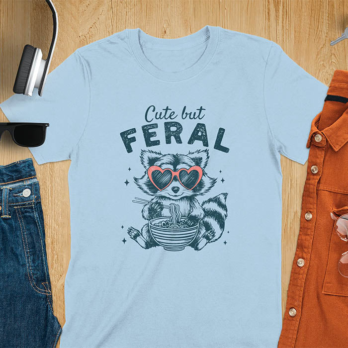 A t-shirt with a raccoon in heart sunglasses eating ramen. Text: "Cute but FERAL". A Funny Cute But Feral Raccoon Shirt, Vintage Retro Graphic Ramen Tee, Silly Sarcastic Tshirt, Hilarious Gag Gift. Unhinged, trash panda, trendy design. Features kawaiiness cuteness. Great gift for animal person, raccoon lover, mom, feral mom. Vintage inspired Animal Lover T-shirt, almost feral shirt.