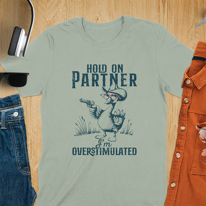 A t-shirt with a goose wearing a cowboy hat and holding a pistol. Text: "HOLD ON PARTNER" above and "I'm OVERSTIMULATED" below. This cowboy frog shirt is a funny meme shirt and oddly specific t shirt. A retro graphic tee and unhinged tshirt, a funny animal shirt. An animal person gift or goose lovers gift, an animal lover gag or mom gift for overstimulated memes.