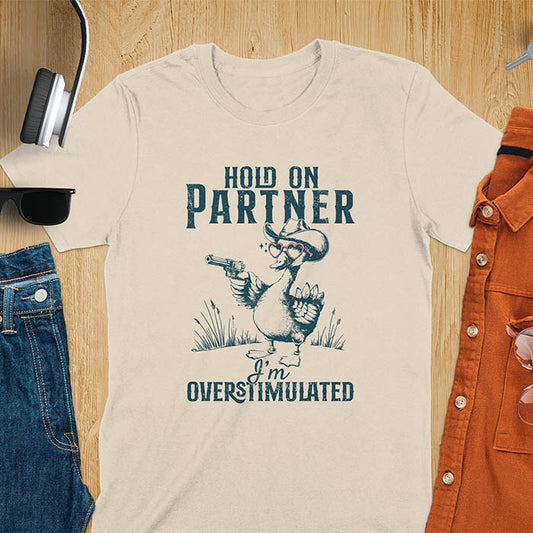 A t-shirt with a goose wearing a cowboy hat and holding a pistol. Text: "HOLD ON PARTNER" above and "I'm OVERSTIMULATED" below. This cowboy frog shirt is a funny meme shirt and oddly specific t shirt. A retro graphic tee and unhinged tshirt, a funny animal shirt. An animal person gift or goose lovers gift, an animal lover gag or mom gift for overstimulated memes.