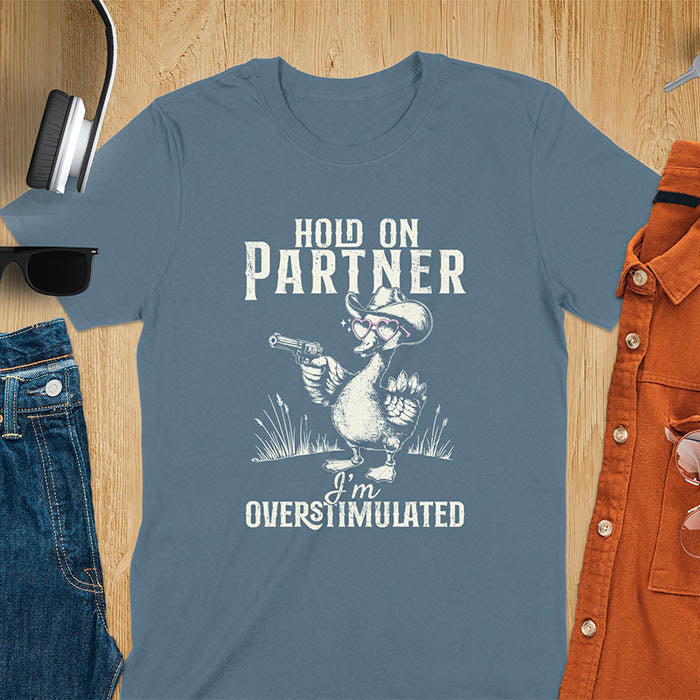 A t-shirt with a goose wearing a cowboy hat and holding a pistol. Text: "HOLD ON PARTNER" above and "I'm OVERSTIMULATED" below. This cowboy frog shirt is a funny meme shirt and oddly specific t shirt. A retro graphic tee and unhinged tshirt, a funny animal shirt. An animal person gift or goose lovers gift, an animal lover gag or mom gift for overstimulated memes.