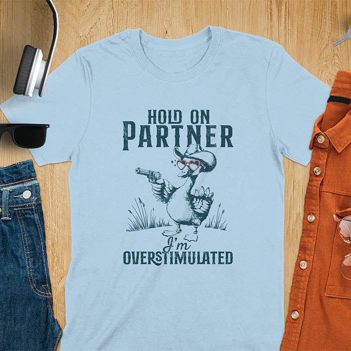 A t-shirt with a goose wearing a cowboy hat and holding a pistol. Text: "HOLD ON PARTNER" above and "I'm OVERSTIMULATED" below. This cowboy frog shirt is a funny meme shirt and oddly specific t shirt. A retro graphic tee and unhinged tshirt, a funny animal shirt. An animal person gift or goose lovers gift, an animal lover gag or mom gift for overstimulated memes.