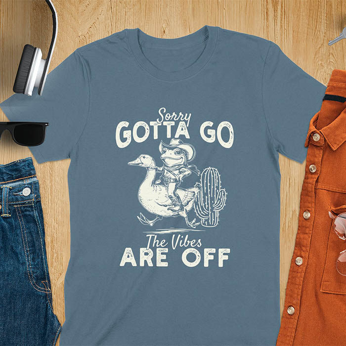 A t-shirt with a frog in cowboy attire riding a goose. Text: "Sorry GOTTA GO" above and "The Vibes ARE OFF" below. This humorous tee-shirt features a funny frog tee and silly funny goose, a weird graphics shirt. A sarcastic meme shirt and funny genz meme, with rodeo cowboy and cowgirl t shirt elements. A vintage graphic tee and western style, an animal person gift. A funny unhinge shirt or mom gift alternative.