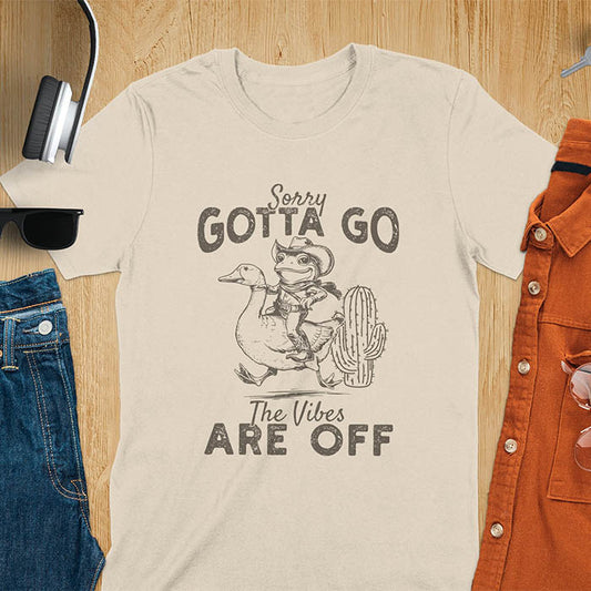 A t-shirt with a frog in cowboy attire riding a goose. Text: "Sorry GOTTA GO" above and "The Vibes ARE OFF" below. This humorous tee-shirt features a funny frog tee and silly funny goose, a weird graphics shirt. A sarcastic meme shirt and funny genz meme, with rodeo cowboy and cowgirl t shirt elements. A vintage graphic tee and western style, an animal person gift. A funny unhinge shirt or mom gift alternative.