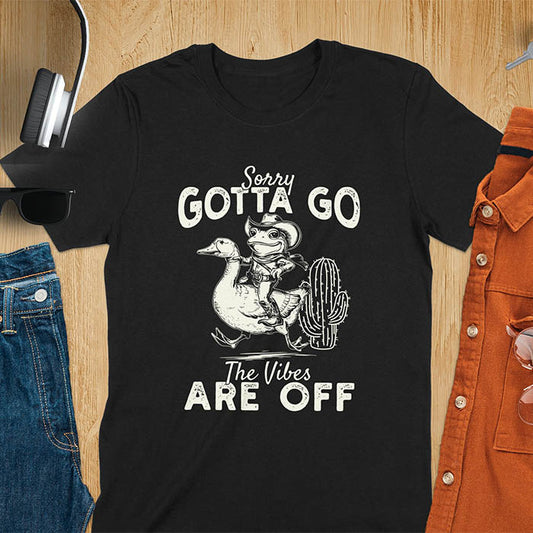 A t-shirt with a frog in cowboy attire riding a goose. Text: "Sorry GOTTA GO" above and "The Vibes ARE OFF" below. This humorous tee-shirt features a funny frog tee and silly funny goose, a weird graphics shirt. A sarcastic meme shirt and funny genz meme, with rodeo cowboy and cowgirl t shirt elements. A vintage graphic tee and western style, an animal person gift. A funny unhinge shirt or mom gift alternative.