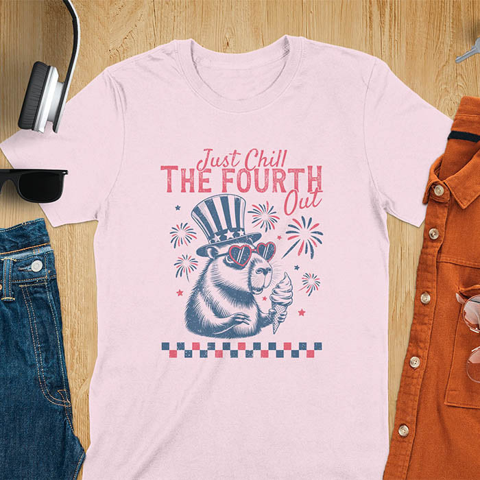 A t-shirt with the text 'Just Chill The Fourth Out' in red and blue lettering. A cartoon illustration of a capybara wearing a red, white, and blue top hat and sunglasses and holding an ice cream cone is below the text. The background of the illustration is a red, white, and blue fireworks display.