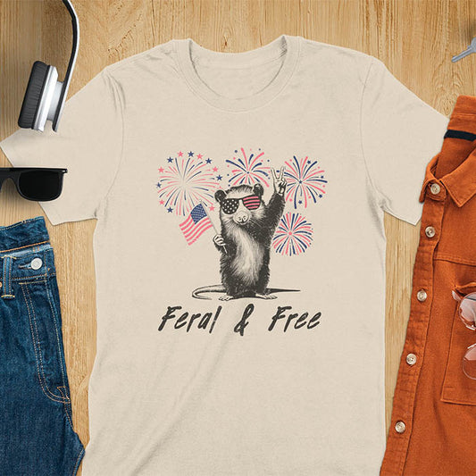 A t-shirt with an opossum wearing patriotic sunglasses. It holds a flag & makes a peace sign, with fireworks behind. Text: "Feral & Free". This unhinged meme tshirt is a merica shirt for Funny 4th of July. A summer tee-shirt with American pride shirt & feral tee-shirt vibes. Features opossum & animal cuteness tee, a feral raccoon shirt for USA Independence Day. A retro america t shirt as an animal person gift or gift for mom.
