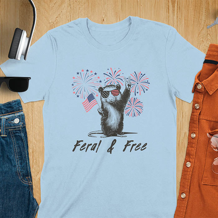 A t-shirt with an opossum wearing patriotic sunglasses. It holds a flag & makes a peace sign, with fireworks behind. Text: "Feral & Free". This unhinged meme tshirt is a merica shirt for Funny 4th of July. A summer tee-shirt with American pride shirt & feral tee-shirt vibes. Features opossum & animal cuteness tee, a feral raccoon shirt for USA Independence Day. A retro america t shirt as an animal person gift or gift for mom.