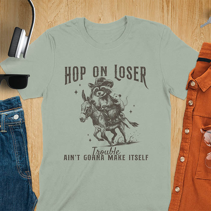 A raccoon riding a donkey with an opossum clinging to its back. Text reads Hop On Loser and Trouble Ain't Gonna Make Itself.