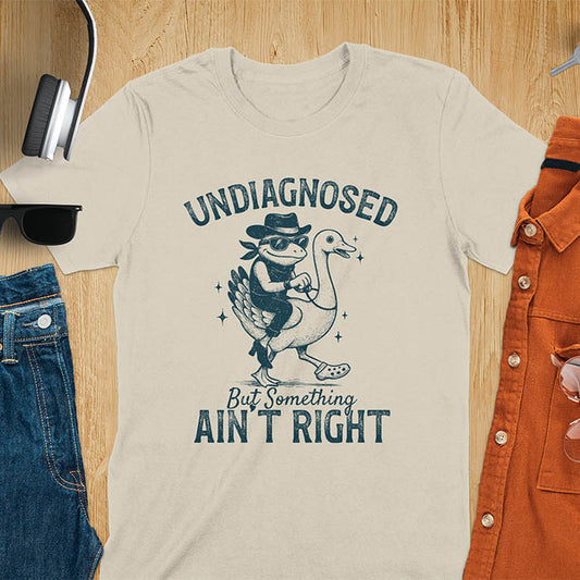 Funny cowboy frog riding goose t-shirt in sand color featuring cartoon frog in cowboy hat and boots riding a goose with text 'UNDIAGNOSED - But Something AIN'T RIGHT