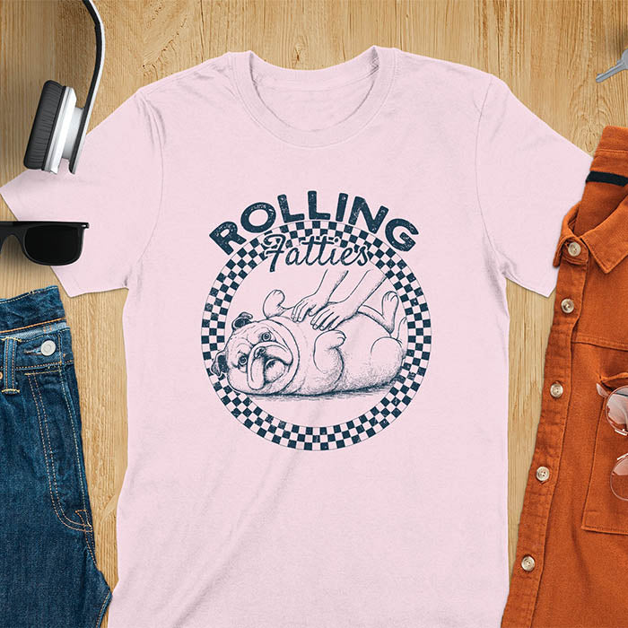 Light pink colored unisex t-shirt featuring 'Rolling Fatties' text in blue lettering above a cute chubby bulldog lying on its back within a blue and white checkered circle border