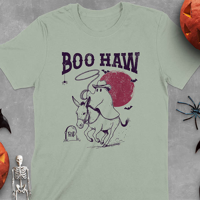 Sage green t-shirt featuring cowboy ghost riding a donkey with lasso, 'BOO HAW' text above, red moon background with bats and RIP tombstone