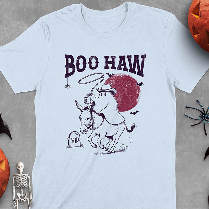 Light blue t-shirt featuring cowboy ghost riding a donkey with lasso, 'BOO HAW' text above, red moon background with bats and RIP tombstone