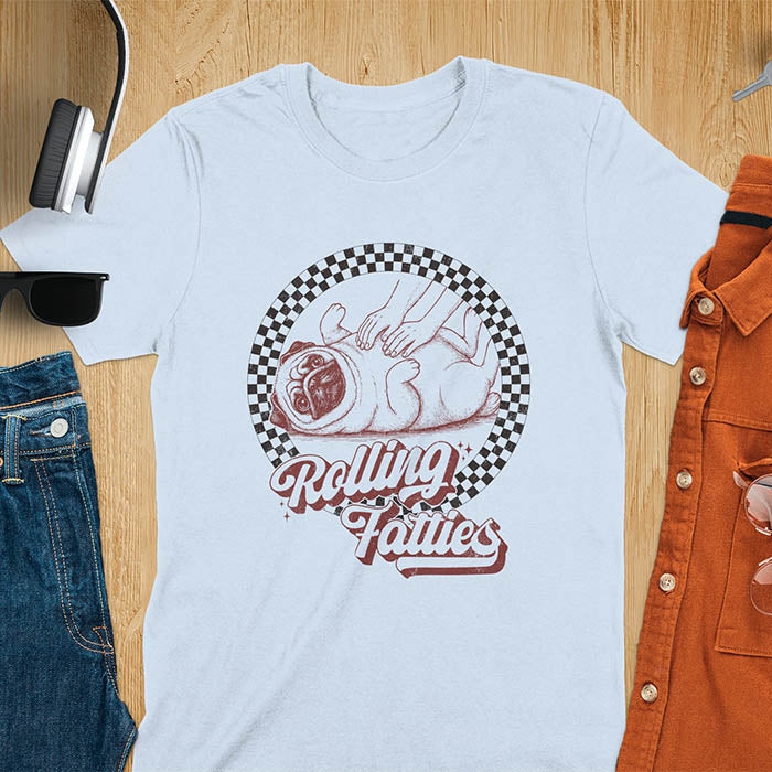 Light blue t-shirt with 'Rolling Fatties' text and illustration of chubby pug lying on its back inside black and white checkered circle border, with human hands reaching toward pug's belly