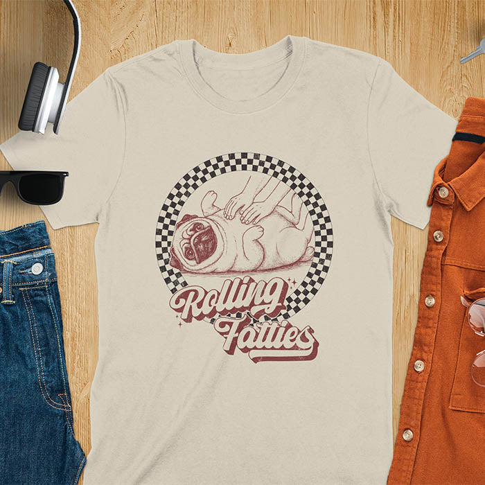 Sand colored t-shirt with 'Rolling Fatties' text and illustration of chubby pug lying on its back inside black and white checkered circle border, with human hands reaching toward pug's belly