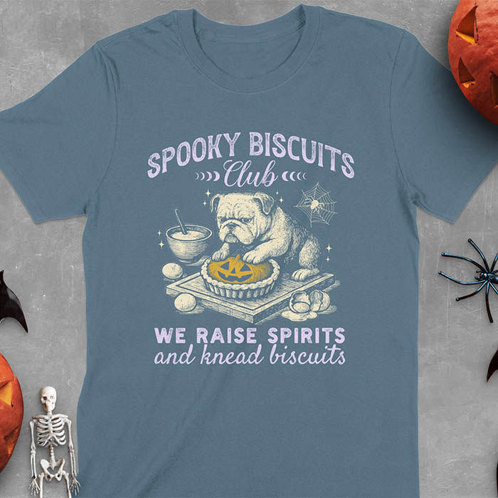 Indigo Blue Light pink t-shirt featuring 'Spooky Biscuits Club' design with illustrated bulldog sitting at wooden table with Halloween pumpkin-shaped biscuit, mixing bowl, eggs, and spider web decoration, text reads 'We raise spirits and knead biscuits'