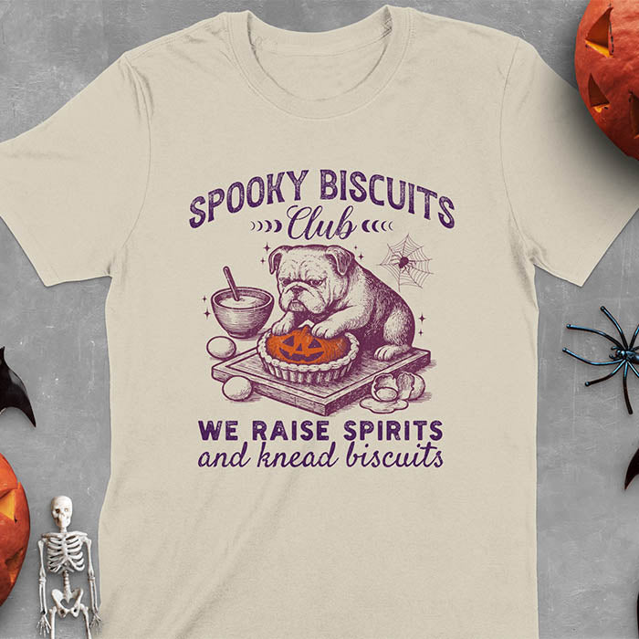 Sand colored t-shirt featuring 'Spooky Biscuits Club' design with illustrated bulldog sitting at wooden table with Halloween pumpkin-shaped biscuit, mixing bowl, eggs, and spider web decoration, text reads 'We raise spirits and knead biscuits'