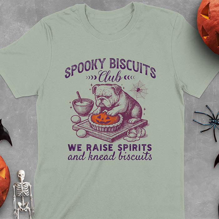 Sage green t-shirt featuring 'Spooky Biscuits Club' design with illustrated bulldog sitting at wooden table with Halloween pumpkin-shaped biscuit, mixing bowl, eggs, and spider web decoration, text reads 'We raise spirits and knead biscuits'