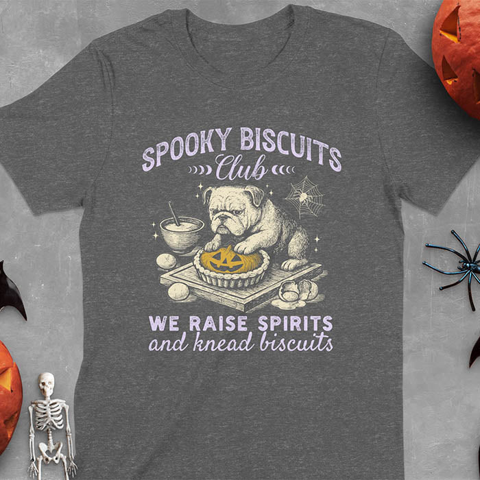 Dark heather t-shirt featuring 'Spooky Biscuits Club' design with illustrated bulldog sitting at wooden table with Halloween pumpkin-shaped biscuit, mixing bowl, eggs, and spider web decoration, text reads 'We raise spirits and knead biscuits'