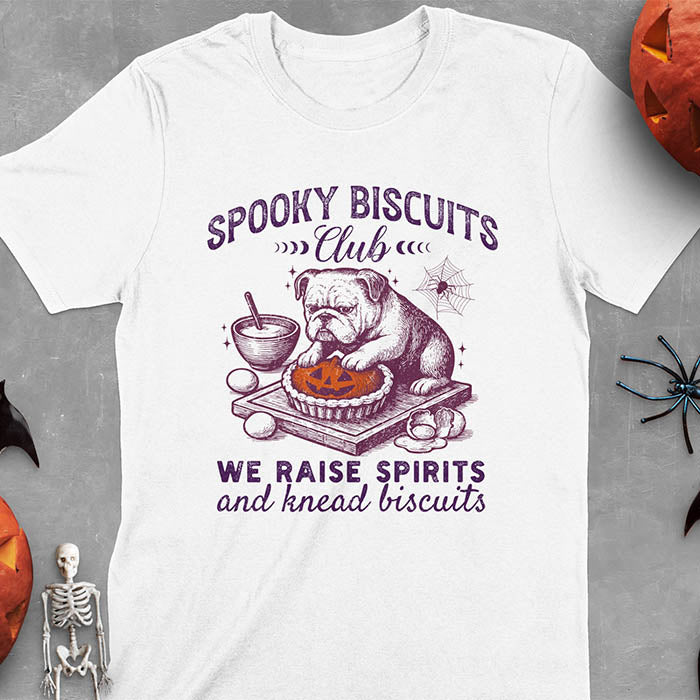 White t-shirt featuring 'Spooky Biscuits Club' design with illustrated bulldog sitting at wooden table with Halloween pumpkin-shaped biscuit, mixing bowl, eggs, and spider web decoration, text reads 'We raise spirits and knead biscuits'