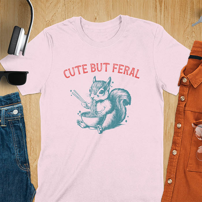 Light pink t-shirt featuring illustrated squirrel holding chopsticks and eating from ramen bowl with red text 'CUTE BUT FERAL' above - playful animal meme design