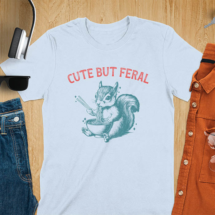 Light blue t-shirt featuring illustrated squirrel holding chopsticks and eating from ramen bowl with red text 'CUTE BUT FERAL' above - playful animal meme design