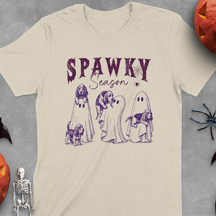 Sand colored Halloween T-shirt featuring 'SPAWKY Season' text with cute ghosts carrying Basset Hound puppies, designed in purple ink with playful Halloween theme