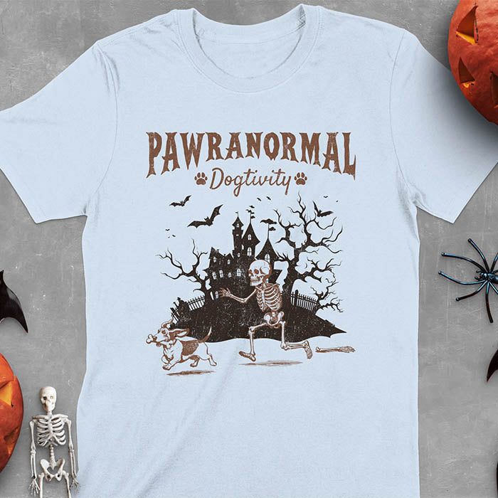 Light blue Halloween T-shirt featuring 'PAWRANORMAL DOGTIVITY' text with humorous scene of Basset Hound running away with skeleton bones while being chased by an angry one-armed skeleton, designed in brown ink with a haunted house background