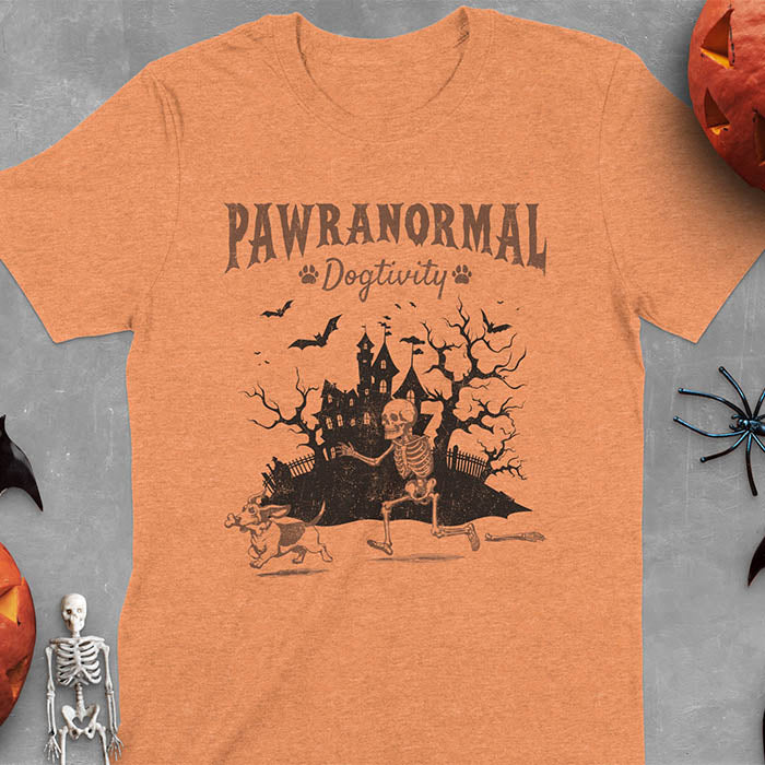 Heather Orange Halloween T-shirt featuring 'PAWRANORMAL DOGTIVITY' text with humorous scene of Basset Hound running away with skeleton bones while being chased by an angry one-armed skeleton, designed in brown ink with a haunted house background