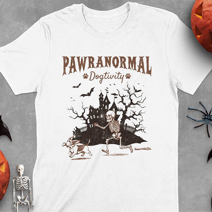 White Halloween T-shirt featuring 'PAWRANORMAL DOGTIVITY' text with humorous scene of Basset Hound running away with skeleton bones while being chased by an angry one-armed skeleton, designed in brown ink with haunted house background