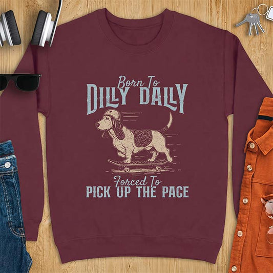 Maroon sweatshirt with a graphic of a basset hound in a skater helmet riding a skateboard with long ears flying back, printed with the text Born To Dilly Dally, Forced To Pick Up The Pace