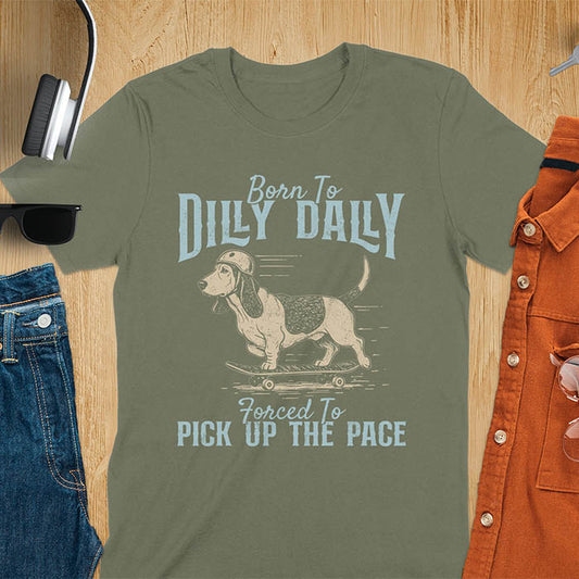 Basset hound wearing helmet on skateboard with Born To Dilly Dally Forced To Pick Up The Pace lettering on military green tee