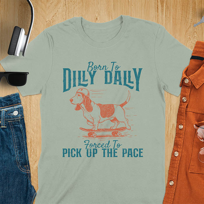 Basset hound wearing helmet on skateboard with Born To Dilly Dally Forced To Pick Up The Pace lettering on sage green tee